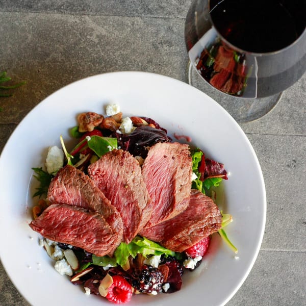 Seared steak slices on a vibrant salad with berries, nuts, and cheese, with a glass of red wine.