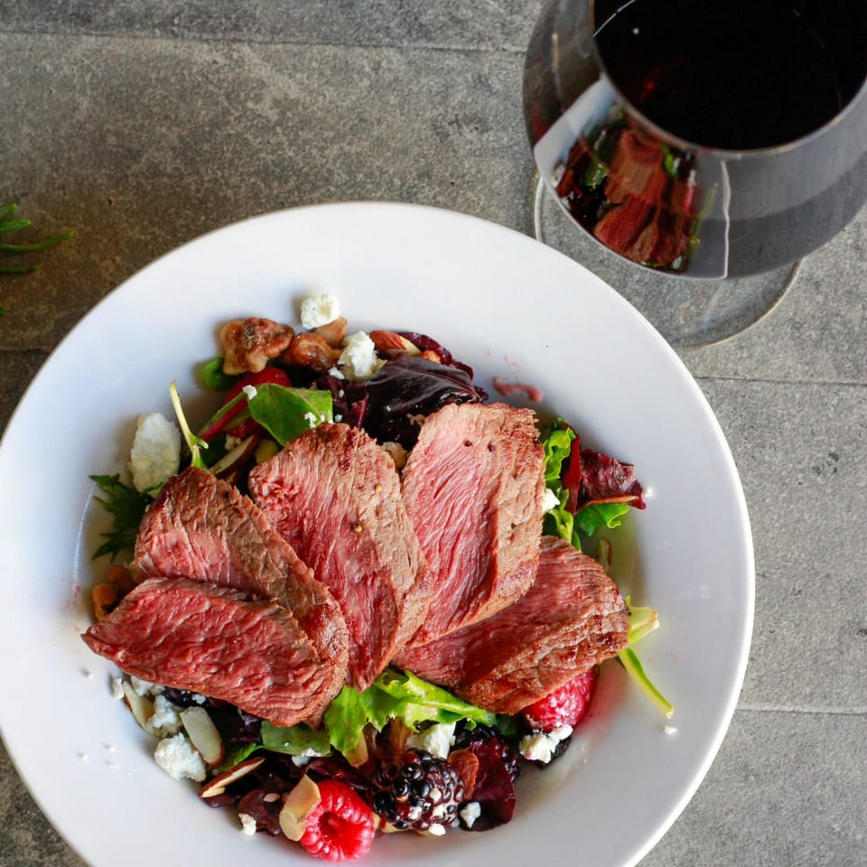 Seared steak slices on a vibrant salad with berries, nuts, and cheese, with a glass of red wine.