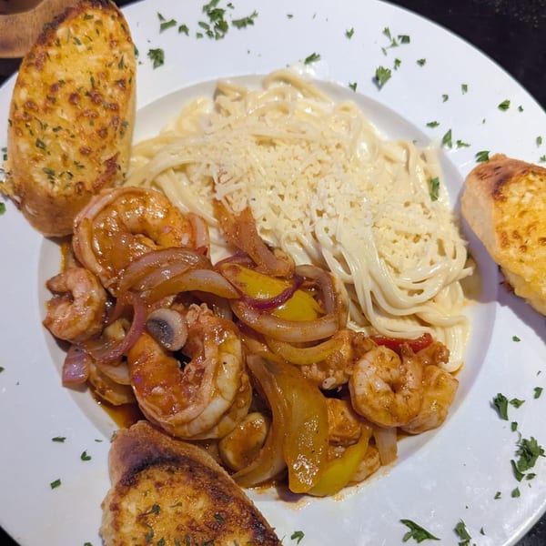 Shrimp scampi with bell peppers and onions, alongside creamy pasta and garlic bread on a white plate.