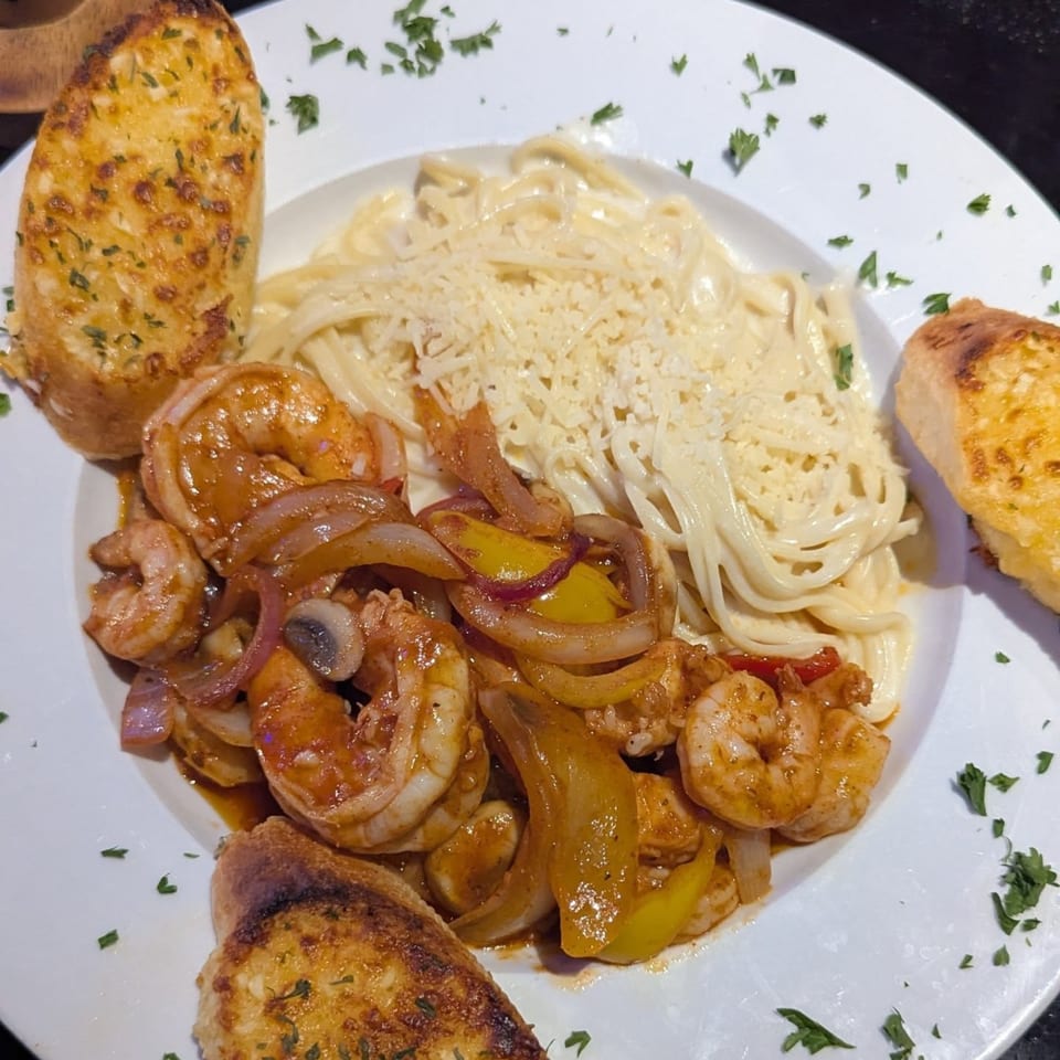 Shrimp scampi with bell peppers and onions, alongside creamy pasta and garlic bread on a white plate.