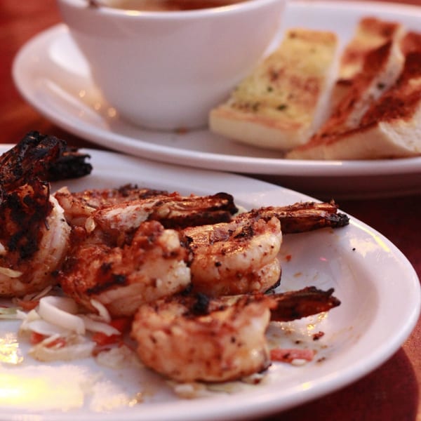 Grilled shrimp with onions and peppers on a white plate, with garlic bread and soup in the background.