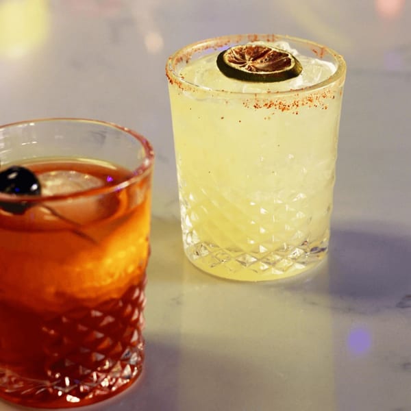 Two cocktails on a bar, one amber with a cherry, the other yellow with a lime.