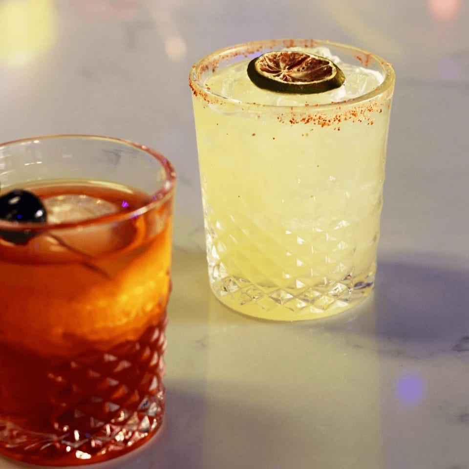 Two cocktails on a bar, one amber with a cherry, the other yellow with a lime.