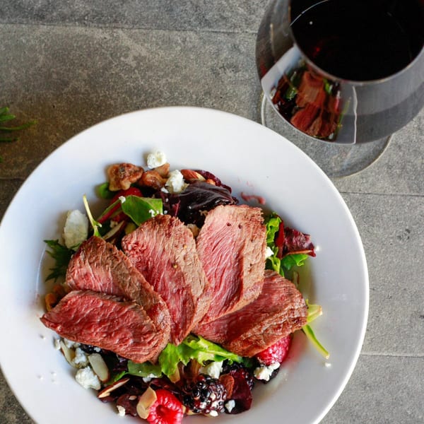 Seared steak slices on a salad with berries, nuts, and cheese, with a glass of red wine.