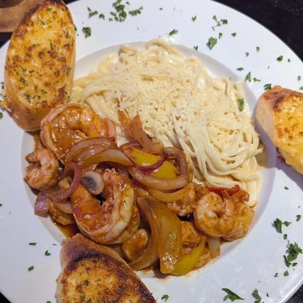 Shrimp scampi with bell peppers and onions, alongside creamy pasta and garlic bread on a white plate.