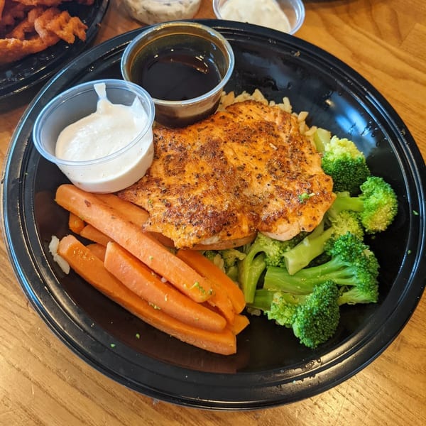 Seasoned salmon with rice, steamed broccoli, carrots, and two dipping sauces in a black bowl.