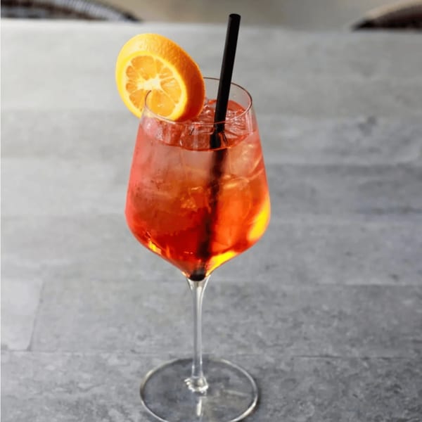 A vibrant orange cocktail with ice, an orange slice, and a black straw in a stemmed glass.