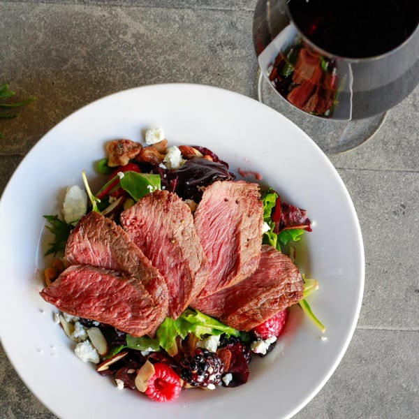 Seared steak slices on a fresh salad with berries, nuts, and feta cheese, with a glass of red wine.