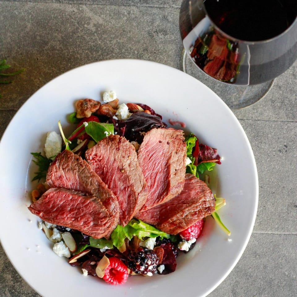 Seared steak slices on a fresh salad with berries, nuts, and feta cheese, with a glass of red wine.