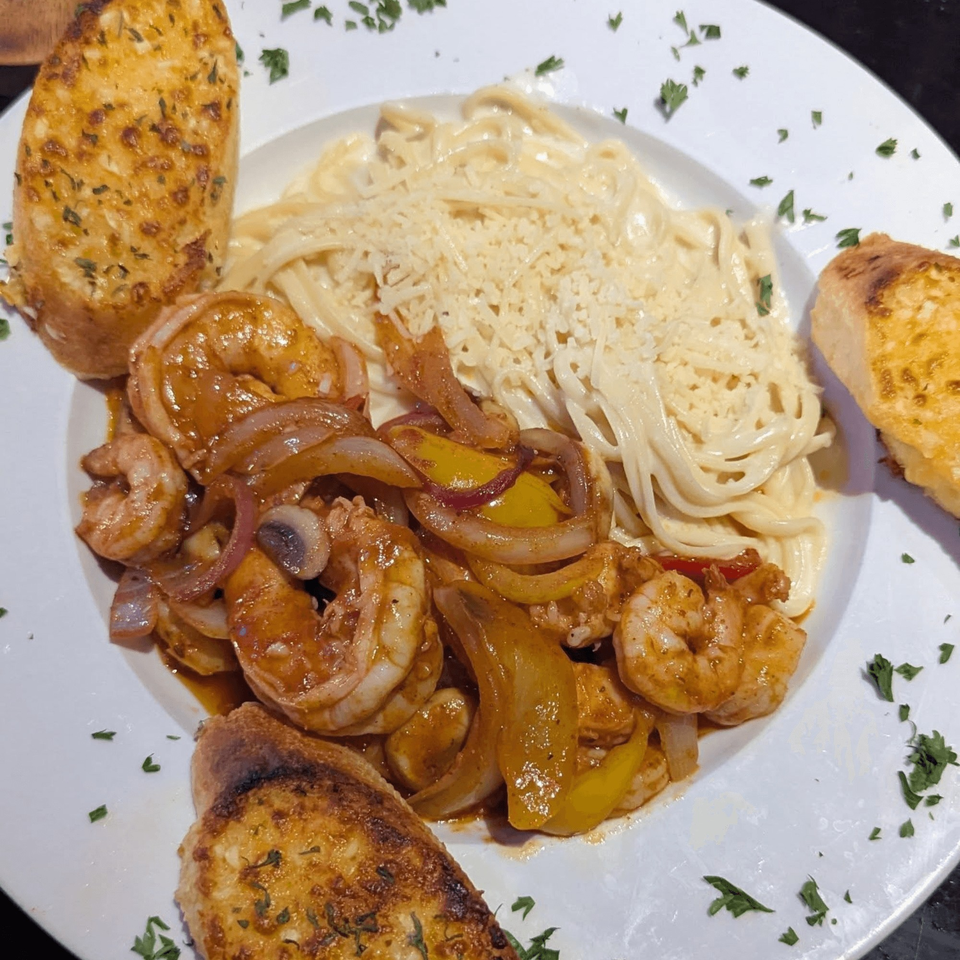 Shrimp scampi with bell peppers and onions, pasta with cheese, and garlic bread on a white plate.