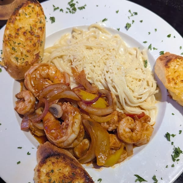 Shrimp scampi with creamy pasta, bell peppers, onions, and garlic bread on a white plate.