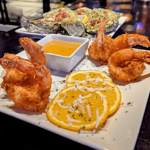 Crispy coconut shrimp with orange slices and dipping sauce, with baked oysters in the background.