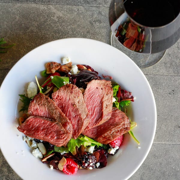 Sliced steak salad with berries, nuts, and cheese, next to a glass of red wine.