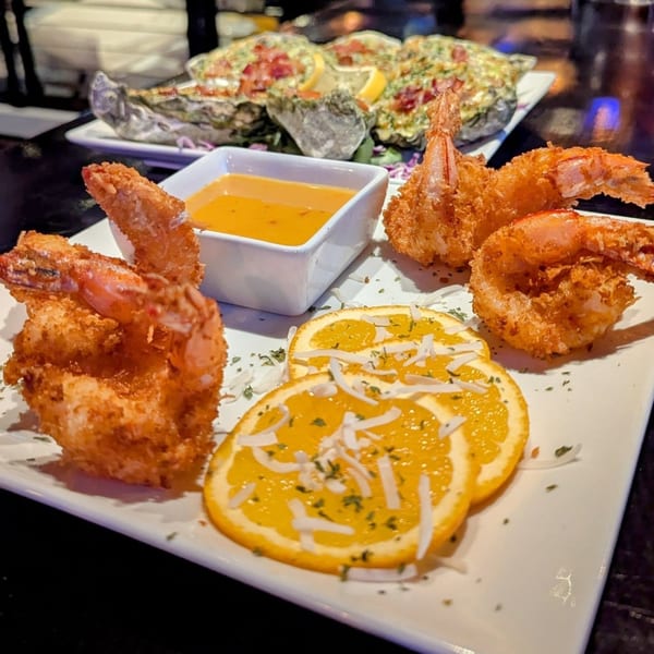 Crispy coconut shrimp with orange slices, dipping sauce, and baked oysters in the background.